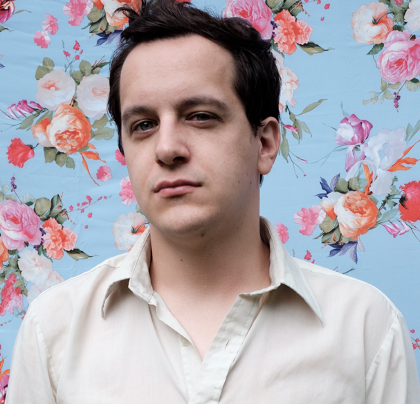 Photo of Ethan Fogus. He wears a cream-colored shirt in front of a multicolored floral background.