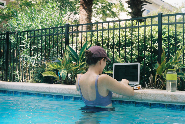 A woman standing in a pool perches her computer on the edge and types.