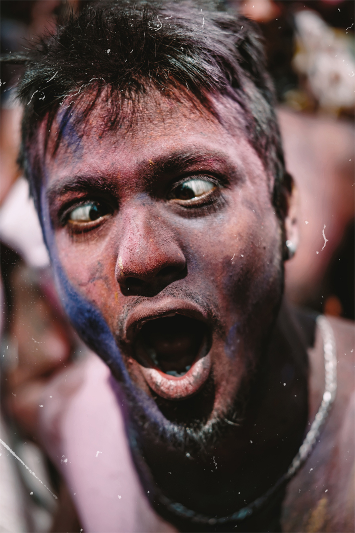 A man covered in multi color pigments is yelling and crossing his eyes.