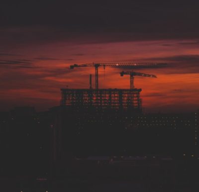 Picture of a large building being constructed with cranes. It is backlit by a red sunset.