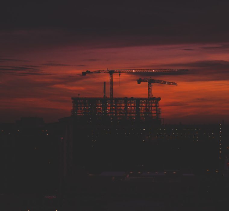 Picture of a large building being constructed with cranes. It is backlit by a red sunset.