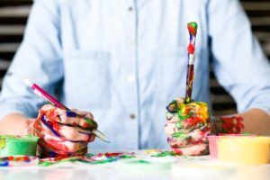 A person holding a paintbrush and pencil. They are covered in multicolor paint.