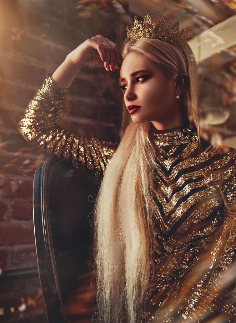 A long-haired woman dressed in a gold dress and crown. She is posing in front of a brick wall.