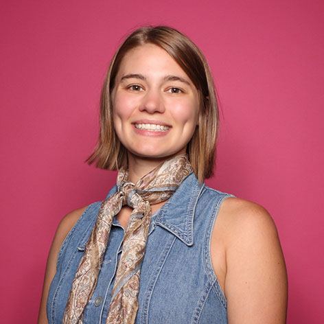 Photo of Belle Newby. She wears a silk scarf and a jean vest, with a pink background.