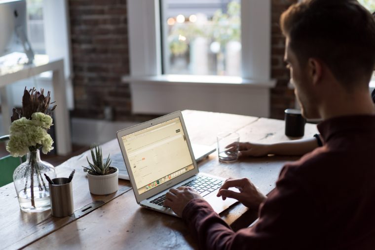 A man typing on a laptop at his desk. There is a person to his right and a succulent on the left.