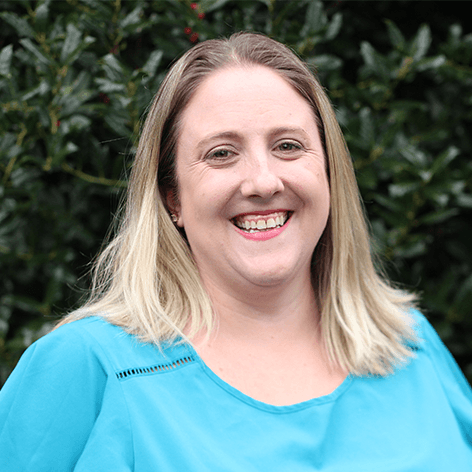 Photo of Caroline Hosey. She wears a blue shirt in front of a green bush.