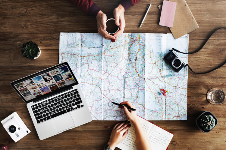 Birdseye view of a laptop, map, and hands. One person draws on map, the other holds a cup of coffee.