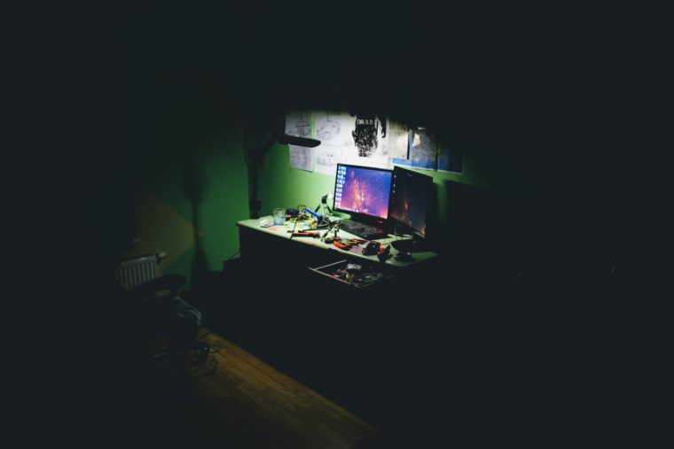 A desk in a dark room. It is lit by a lamp and two monitors. The desk is covered with handtools.