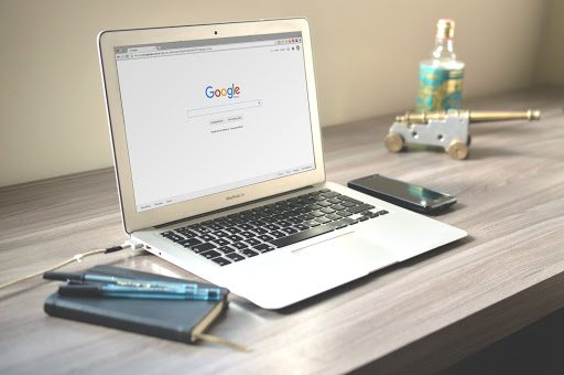 A laptop displaying the Google homepage. There's a phone, journal, pens, and tchotchkes on the desk.