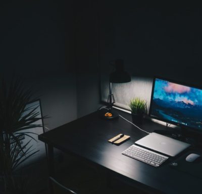 A desk in a dark room. It is lit by a desk lamp and computer monitor.