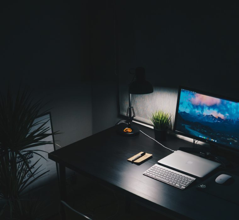 A desk in a dark room. It is lit by a desk lamp and computer monitor.