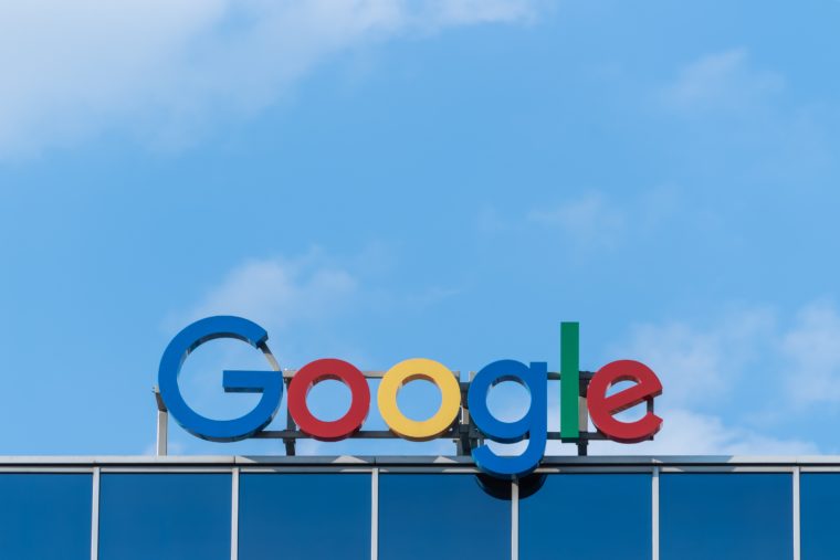 A photo of a Google logo sign on a roof. The sky is blue with white clouds.