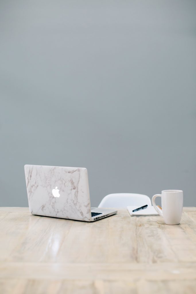 A laptop with a marble case, a pen and journal, and a cup of coffee on a wooden table.