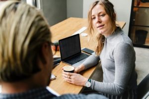 A woman sitting at a desk holding her coffee is talking to a man standing beside her.