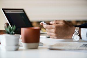 Photo of a man's hands using his phone at a desk. He has a watch, laptop, and some succulents.