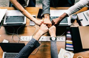 Five people fistbump over a desk with many laptops open.