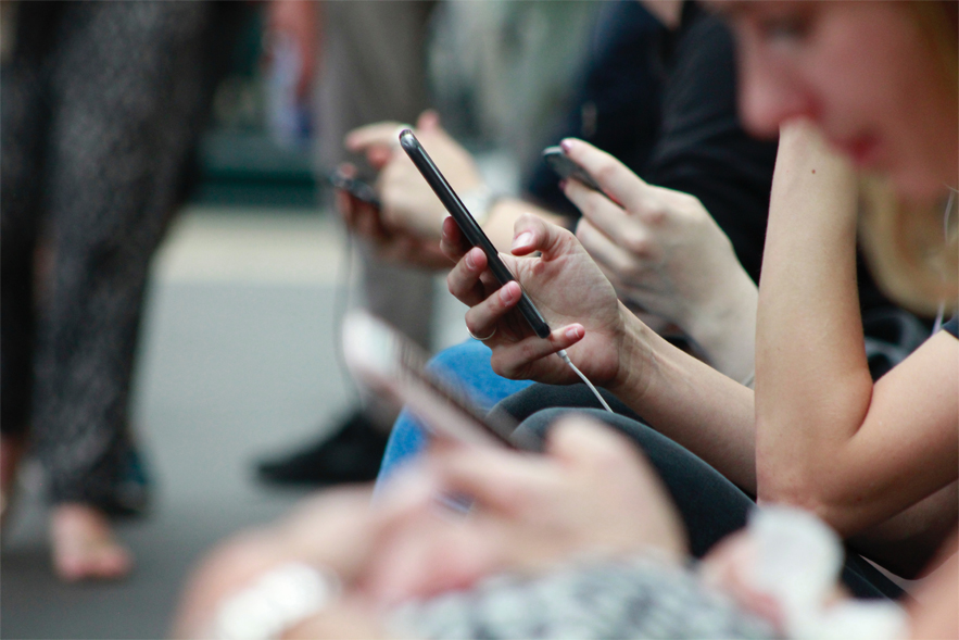 A group of people sitting on a bench outside, all looking at their phones.