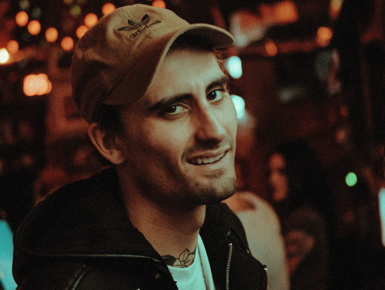 Photo of a man in a tan adidas hat, leather jacket, and white shirt.