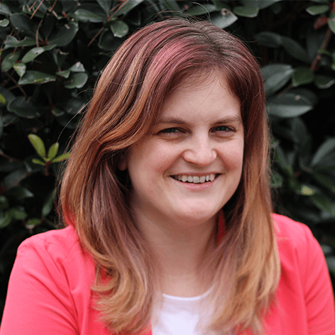 Photo of Stephanie Pietrosanti. She wears a pink sweater in front of a green bush.