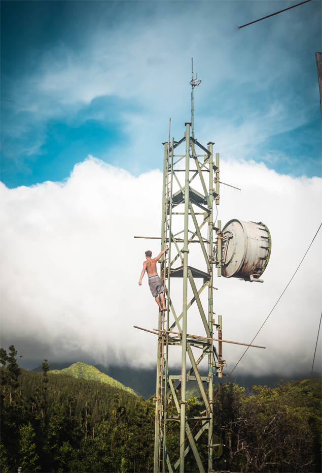 A shirtless man hangs off the top of a water tower looking out over the mountains.