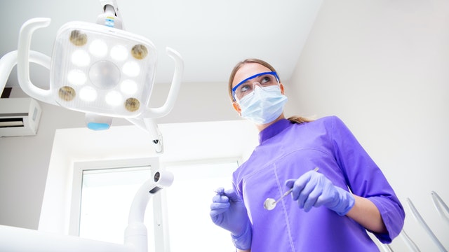 A dental assistant in blue scrubs, a medical mask, and eyeglasses stands above the camera.