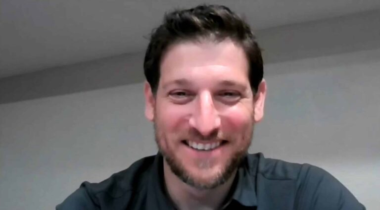 A man smiles at the camera through a computer webcam. He is wearing a gray shirt and has brown hair.
