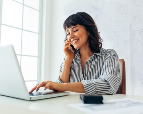 Woman entrepreneur working in office Smiling female architect working on laptop in office while talking over mobile phone. Female entrepreneur sitting at workplace with architecture drawings in the background.